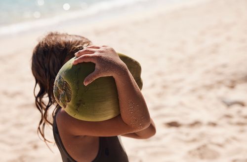 A child walking on the sand at Kokomo Private Island holding a large coconut on her shoulder.