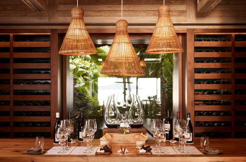 A private dining table within an intimate wine cellar with several large wine glasses set across the table. Several shelves of wine run along the wall separated by a large window looking out into a lush tropical forest.